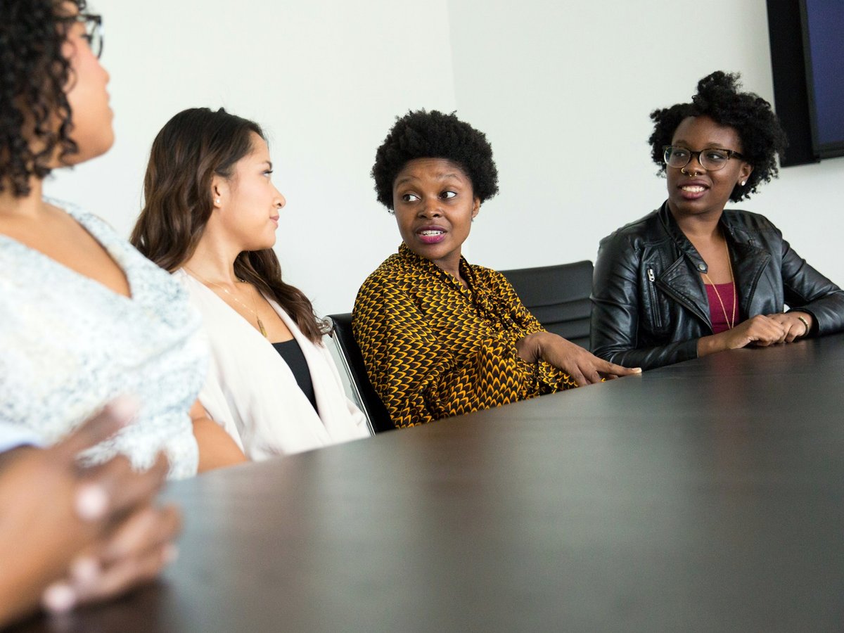 Cinco pessoas sentadas à mesa de uma sala de reunião, conversando e ouvindo umas às outras