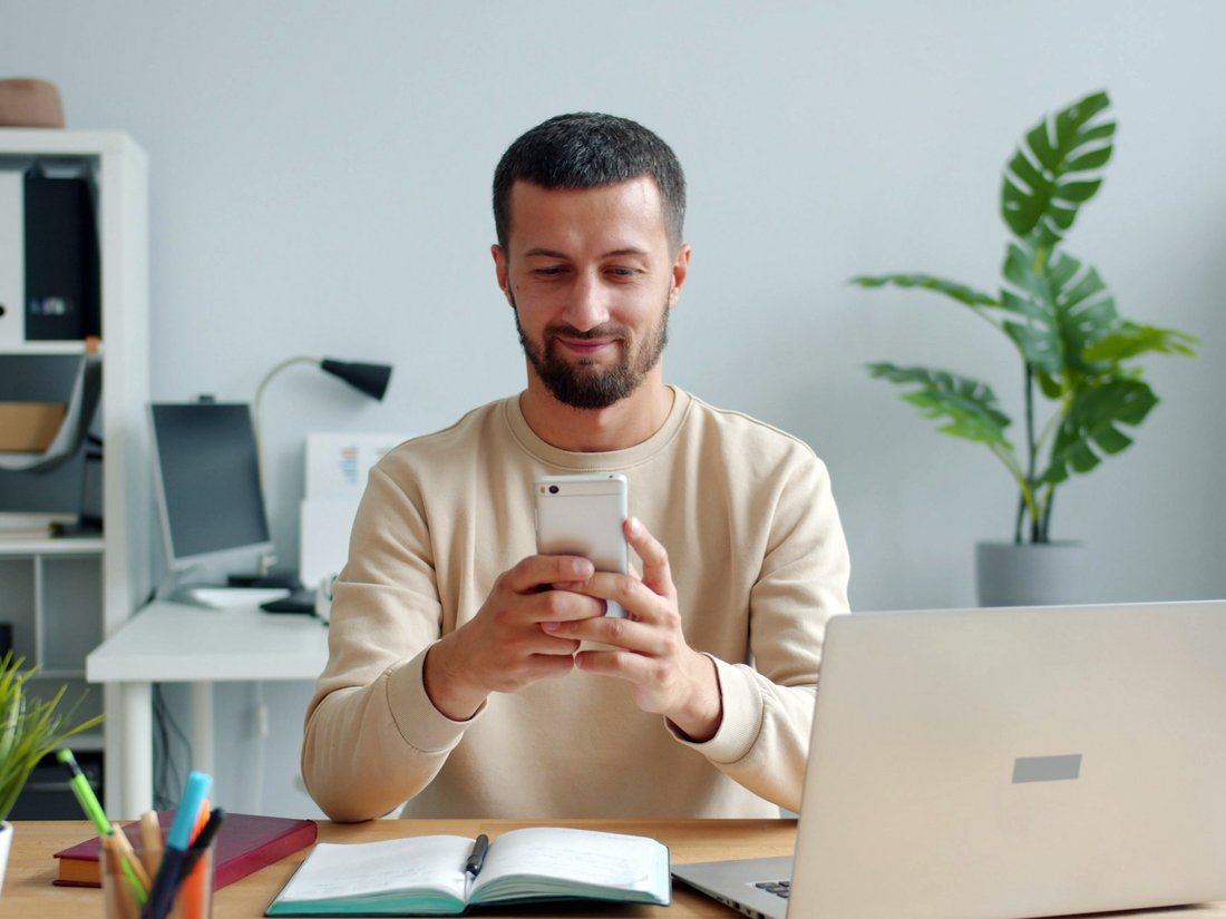 Homem sentado à mesa de trabalho respondendo a um questionário pelo celular, com notebook e caderno abertos