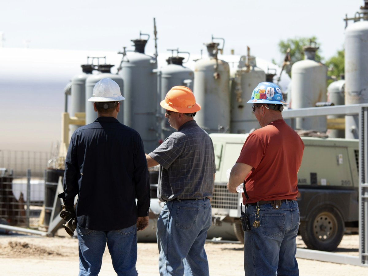 Três profissionais de capacete conversando em uma planta industrial, ao lado de tanques e equipamentos