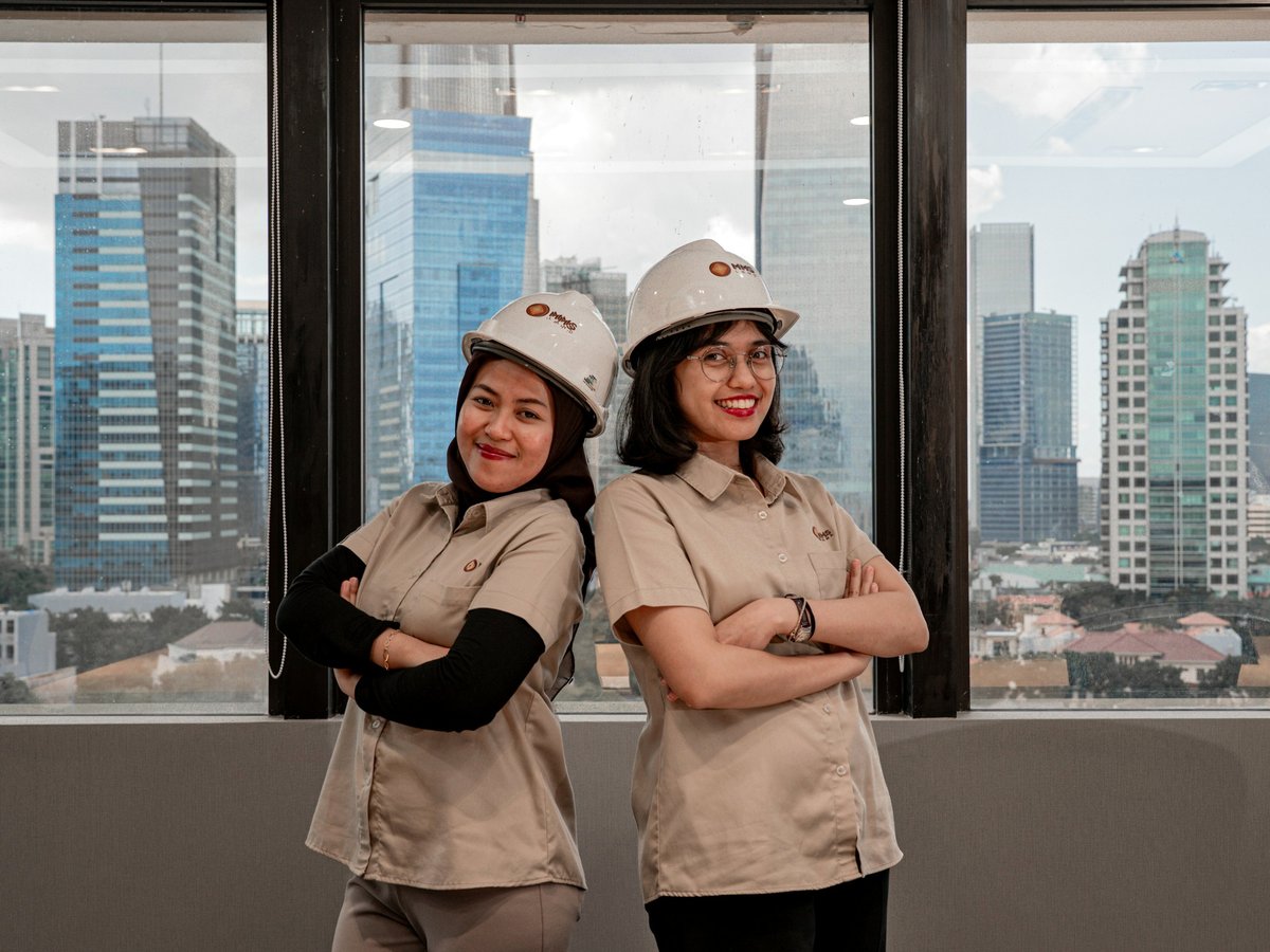 Duas profissionais de capacete e uniforme, de braços cruzados, em um escritório com vista para a cidade