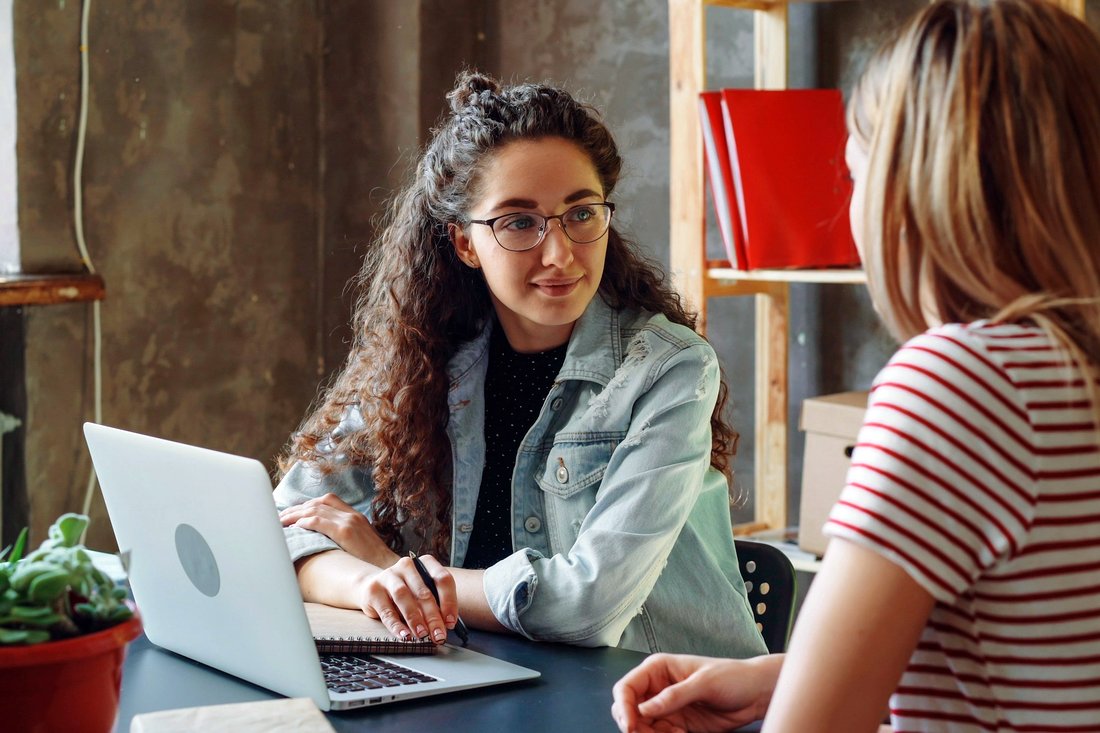 Duas mulheres conversando frente a frente em uma mesa, com um notebook aberto